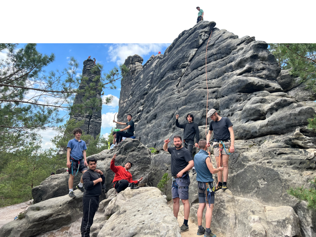 Klettern in der Sächsischen-Schweiz. Mehrere Jungen sitzen und stehen vor einer hohen Felswand und werden für das Klettern vorbereitet, mit Gurten und Seilen. Zufriedene Sieger-Posen und Lächeln.