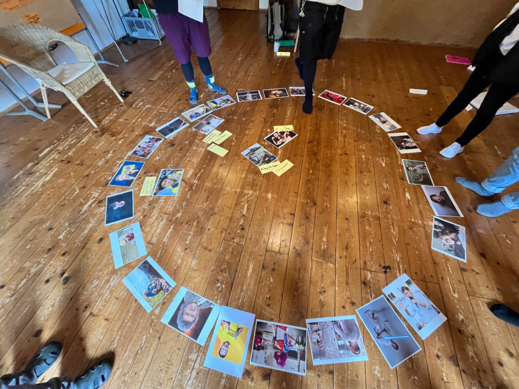 Auf einem Holzfußboden verteilt liegen kreisrund (wie in einer Arena) verschiedene bekannte Portraits von realen und fiktiven Persönlichkeiten. Von den betrachtenden und diskutierenden Personen im Raum sind die Füße und Beine sichtbar.