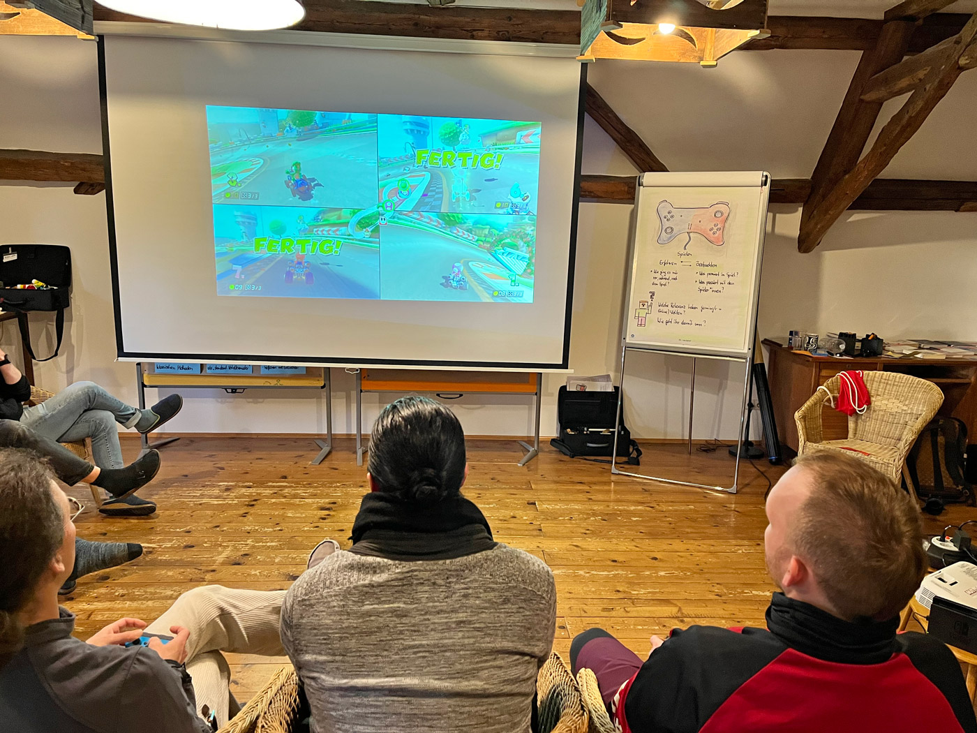 Blick über die Schulter: Mehrere Personen zocken ein Nintendo-Spiel auf einem Splitscreen, projiziert auf eine Leinwand. Im Hintergrund Holzbalken erkennbar, die auf Dachschrägen hinweisen