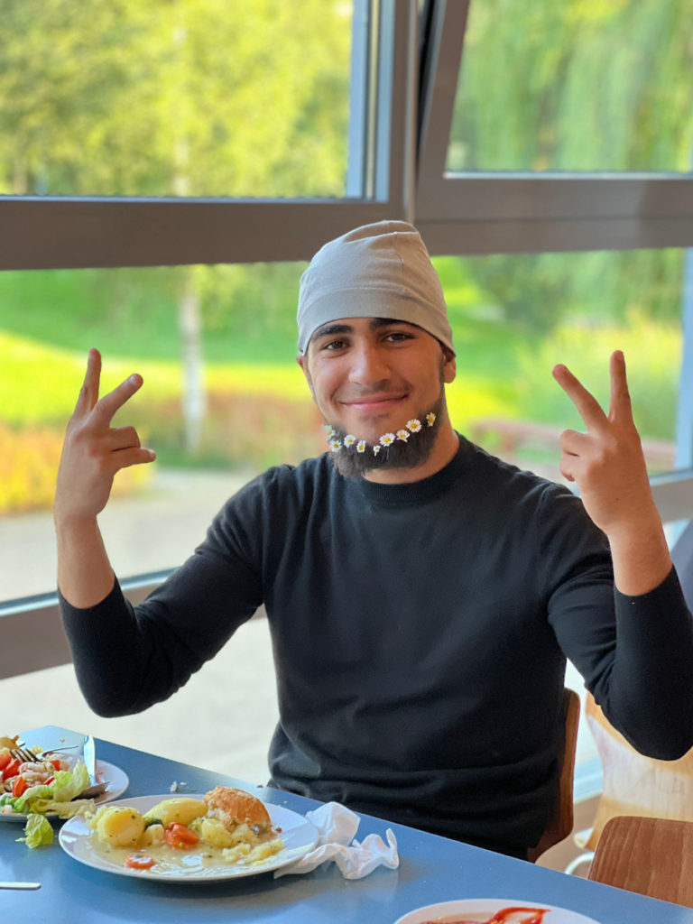 ein fröhlicher Jugendlicher mit Gänseblümchen im Bart, sitzend an einem Tisch, davor ein Teller mit Essen