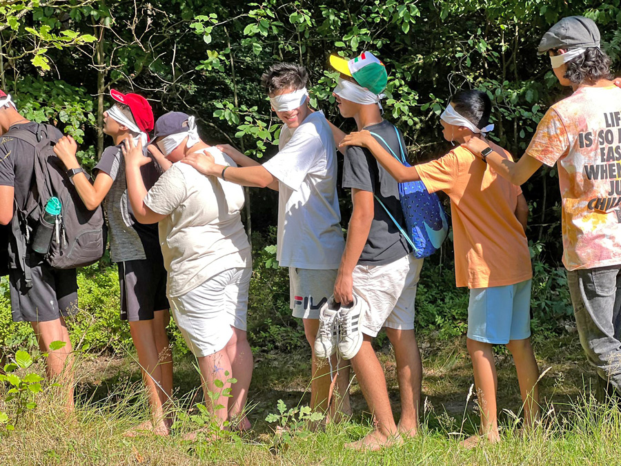 Mehrere Jungen stehen mit verbunden Augen auf einer Wiese hintereinander in Reihe, um ein interaktives Spiel / Methode druchzuführen