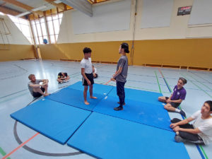 Eine Turnhalle. Im Vordergrund mehrere blaue Matten, darum sitzend ein paar Zuschauende, auf der Matte zwei Jungen, die sich gegenüber stehen vor einem Kampf
